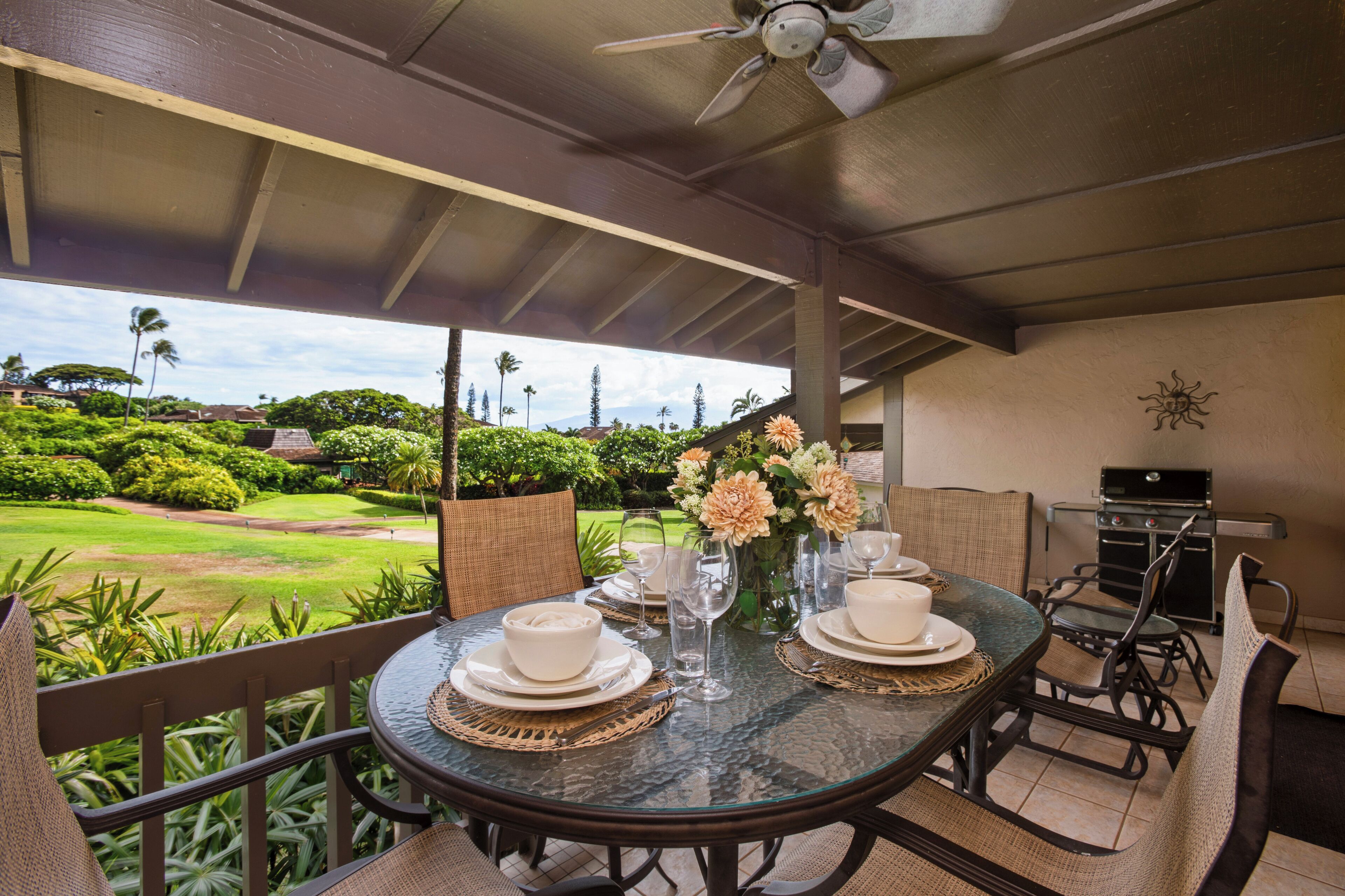 A covered outdoor dining area with a table set for two, surrounded by wicker chairs, a grill, and a view of a golf course.