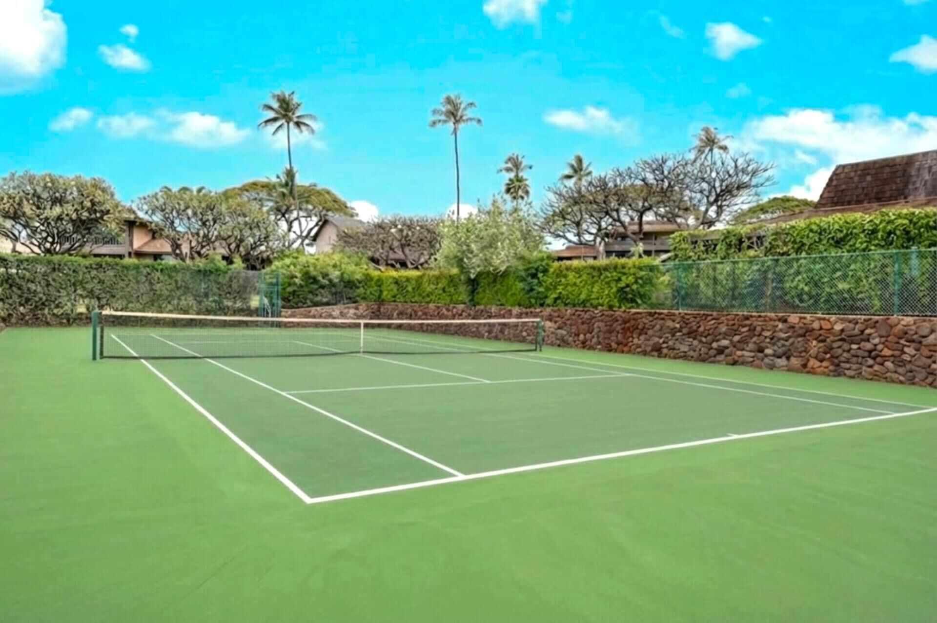 A tennis court with a net, surrounded by greenery and a stone wall.