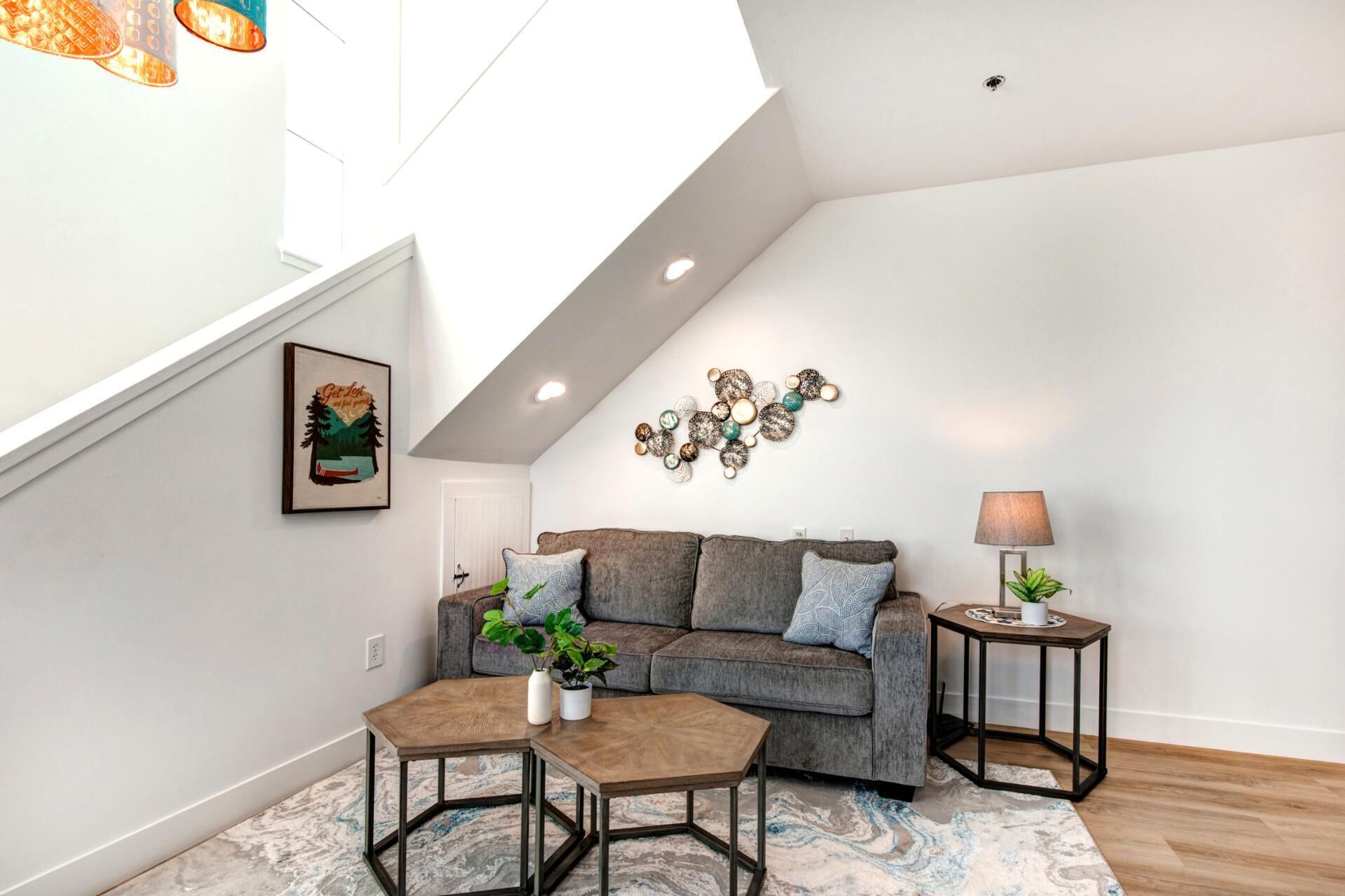 A modern living room with a gray sofa, two matching ottomans, a coffee table, a wall-mounted decorative piece, and a small side table with a lamp and plant.