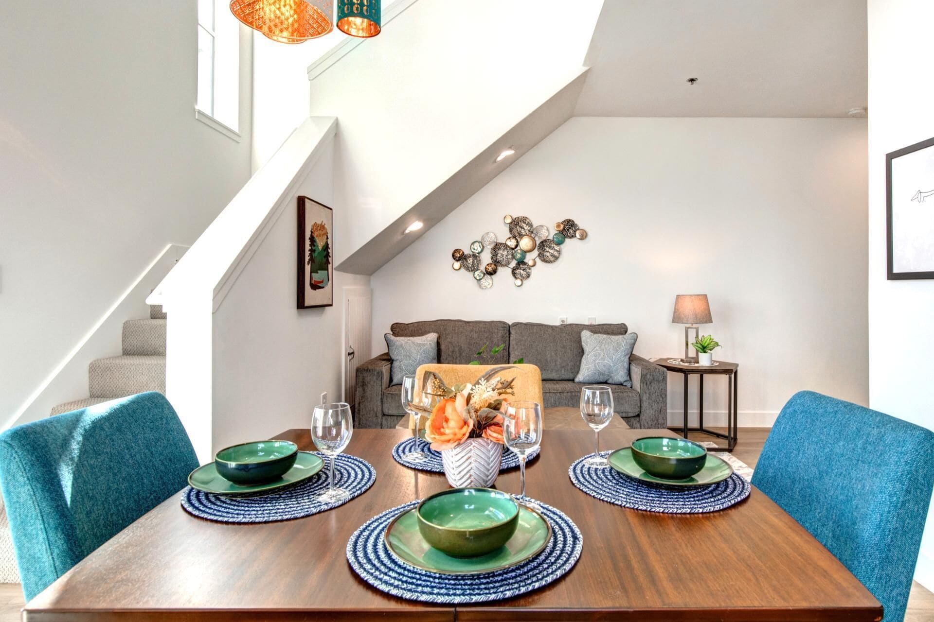 Un coin repas avec une table en bois dressée pour quatre personnes, des chaises bleues et un escalier menant à un niveau supérieur.