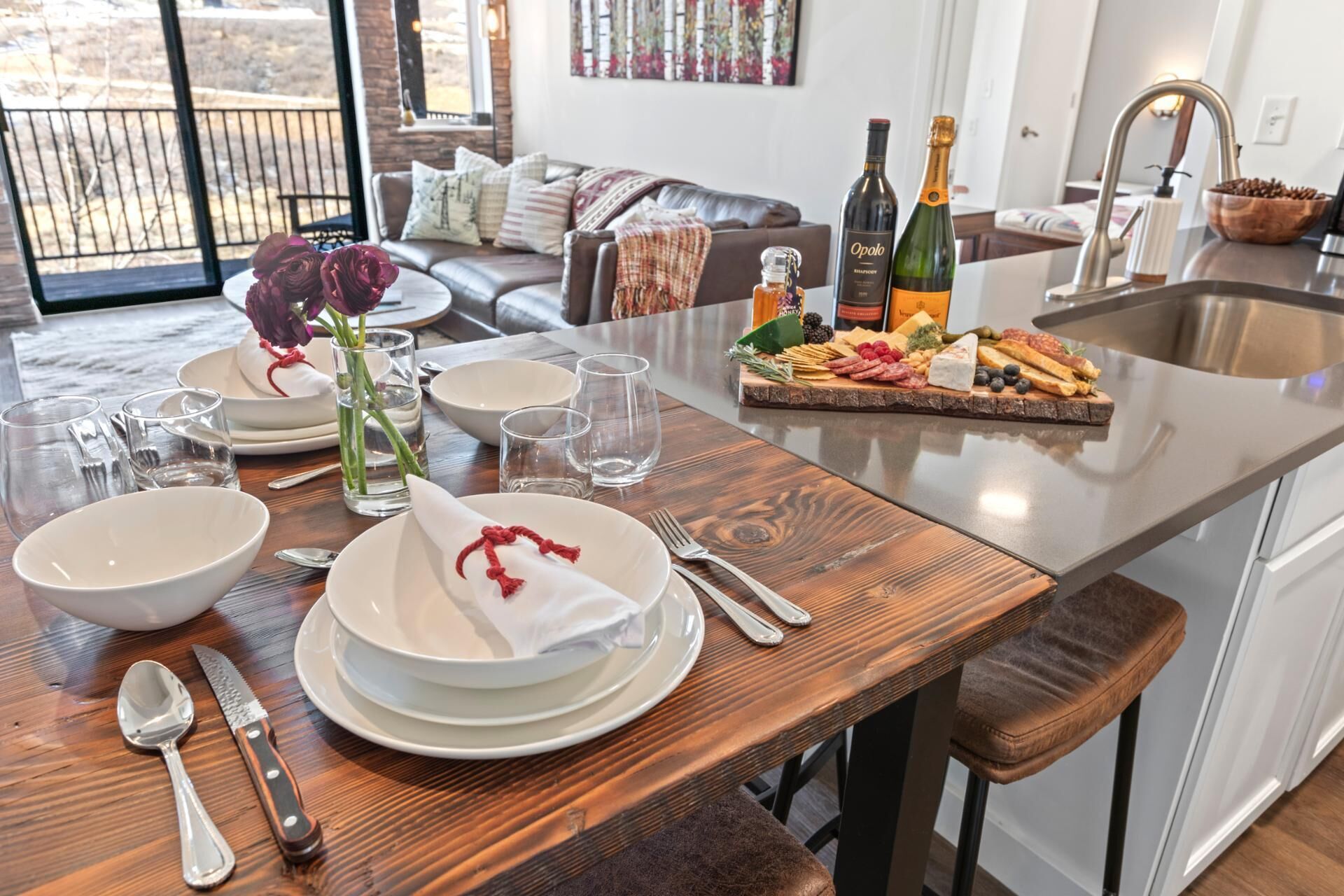 Une cuisine bien éclairée, dotée d’un plan de travail en bois, d’un coin repas avec une table dressée pour quatre personnes, et offrant une vue sur l’extérieur grâce à une grande fenêtre.