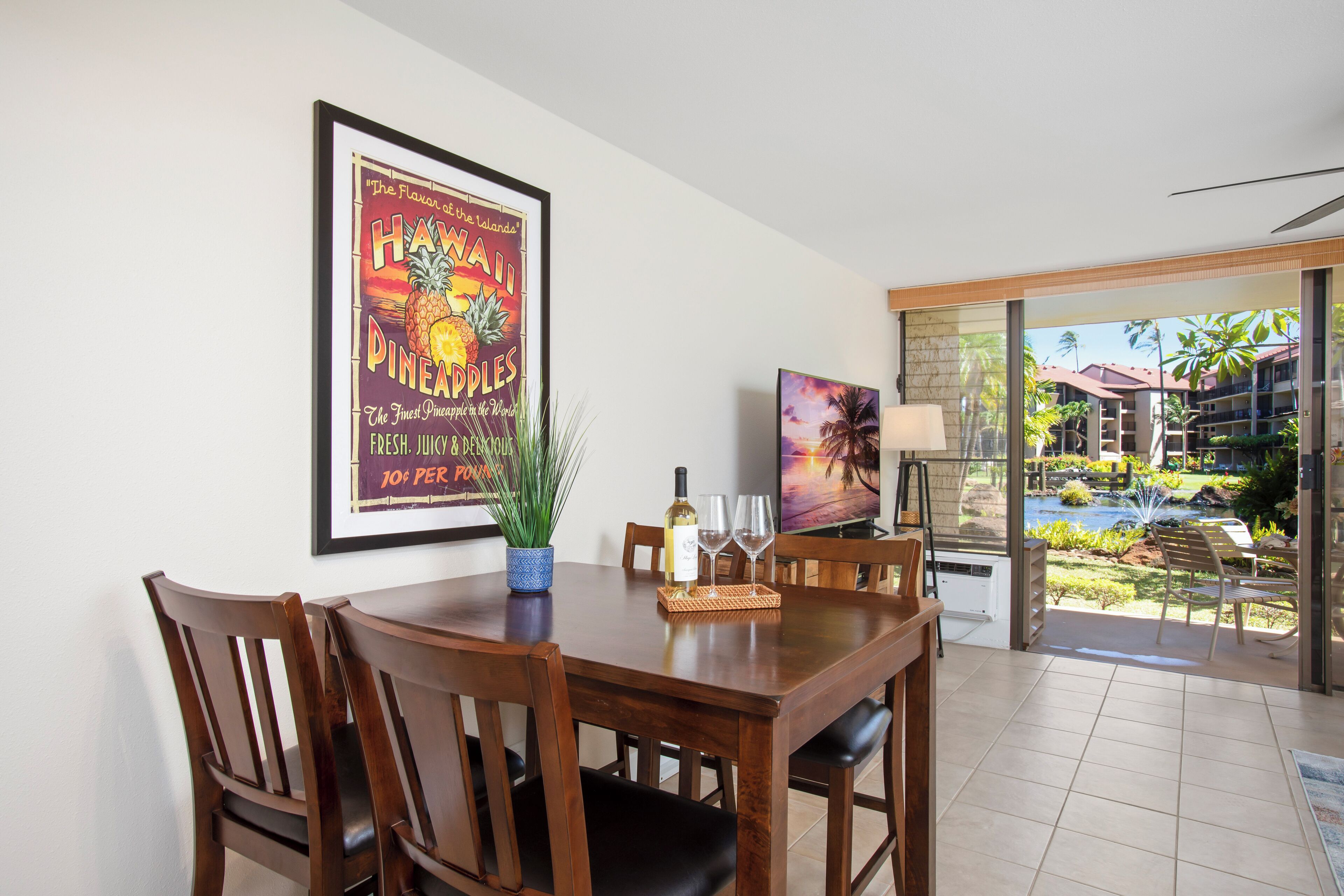 A dining area with a wooden table, chairs, and a framed poster. There is a potted plant, a wine bottle, and glasses on the table. The room has a view of a pool and palm trees.