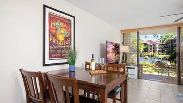 A dining area with a wooden table, chairs, and a framed poster. There is a potted plant, a wine bottle, and glasses on the table. The room has a view of a pool and palm trees.
