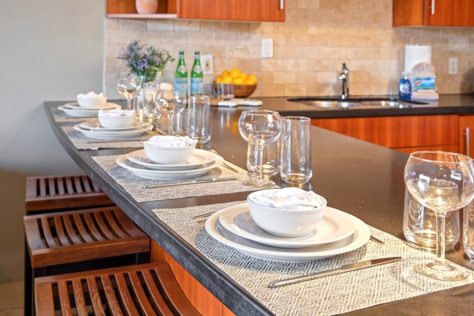 A long dining table set with white plates, bowls, and glasses, with wooden benches on one side.