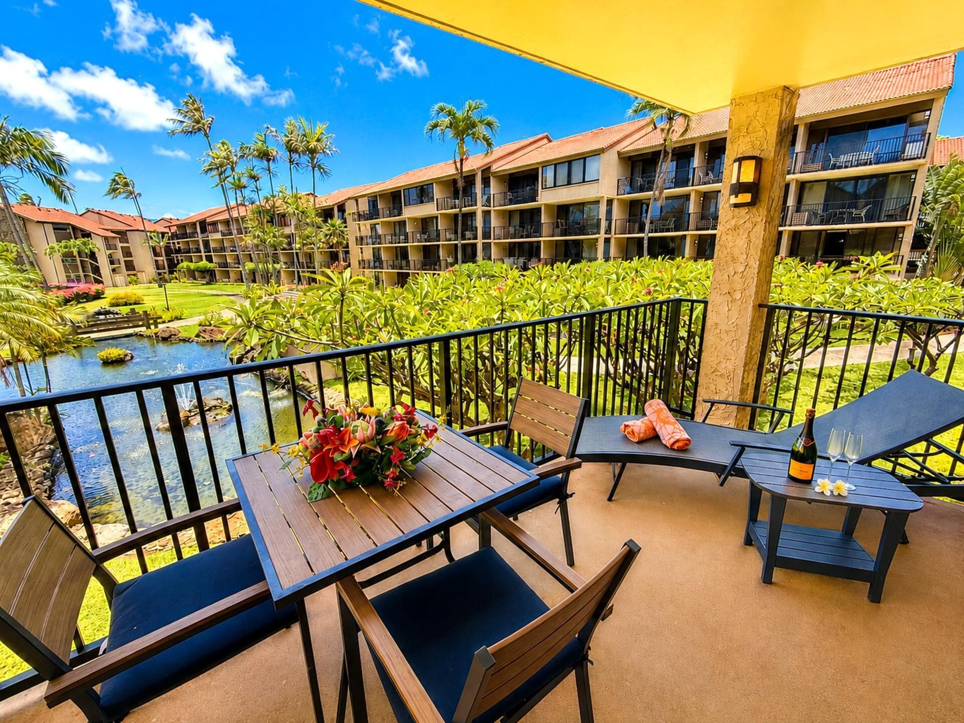 Un balcon d’hôtel avec une table, des chaises, une petite table sur laquelle se trouvent une bouteille et des verres, et une vue sur une piscine et la verdure.