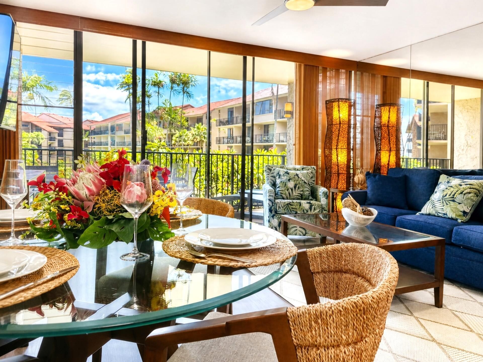 A modern lounge area with a glass table, wicker chairs, a blue sofa, and a balcony with a view of buildings and greenery.