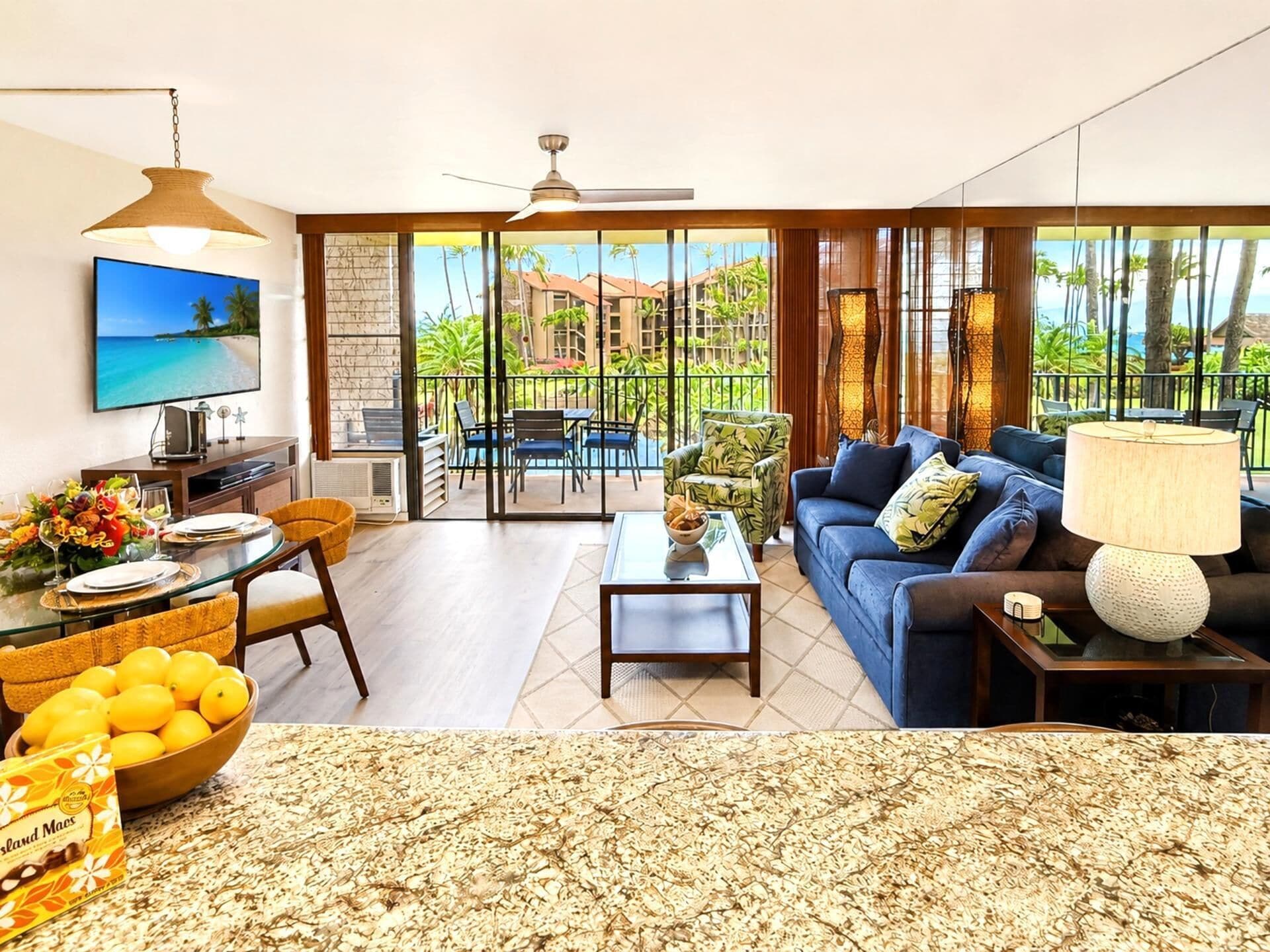 A spacious living room with a large TV, a dining area, and a balcony with a view of palm trees and a building.
