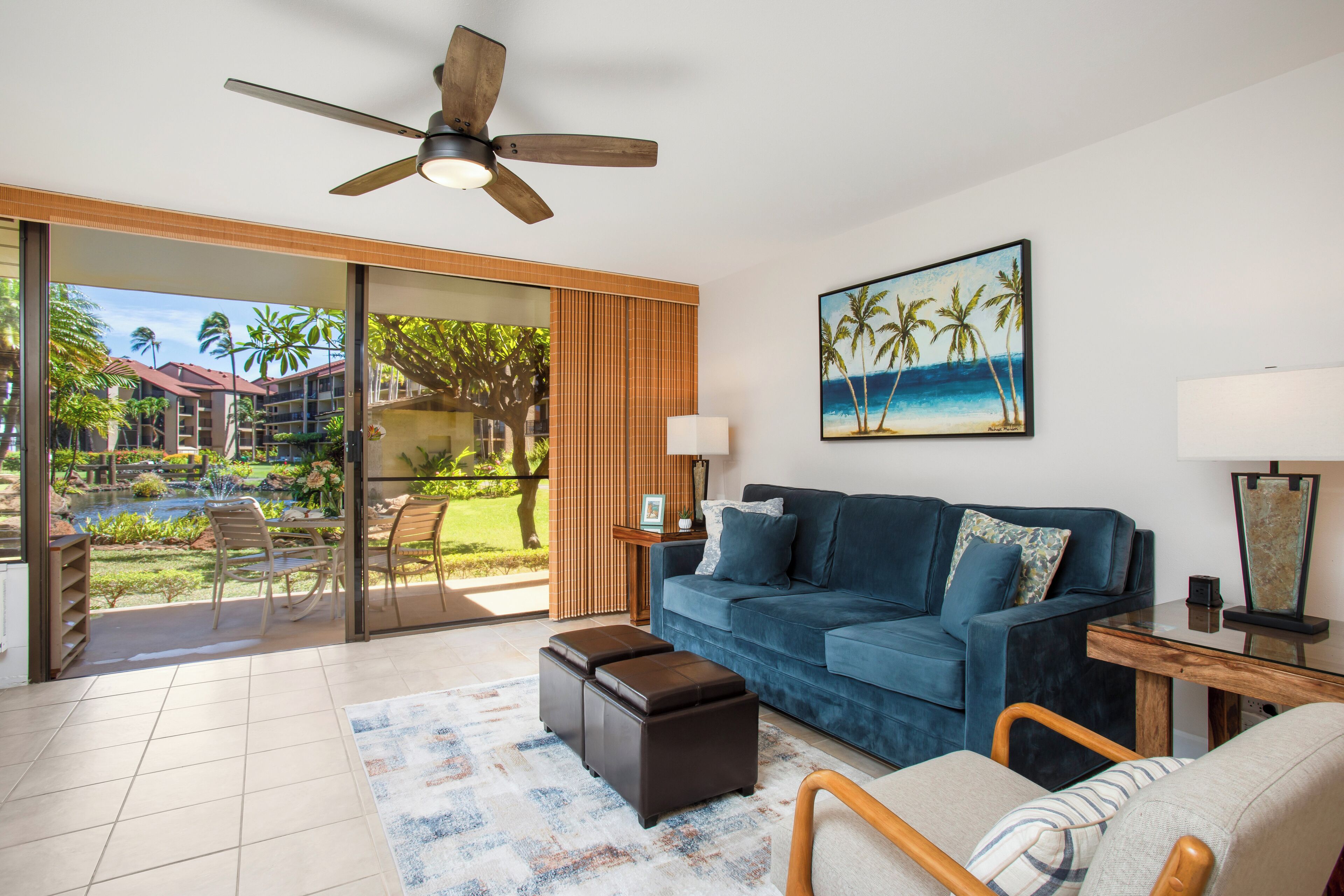 A living room with a blue sofa, wooden coffee table, and a ceiling fan. There is a sliding glass door leading to a balcony with a view of palm trees and a pool.