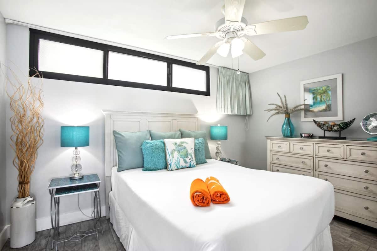 Une chambre à coucher comprenant un lit, deux tables de chevet, une commode, une coiffeuse, un ventilateur de plafond et une fenêtre avec des stores.