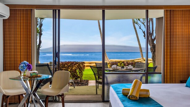 A hotel room with a large sliding glass door leading to a balcony with a view of the ocean and a grassy area.