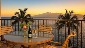 A round table with chairs on a balcony overlooking the sea with palm trees and mountains in the background.