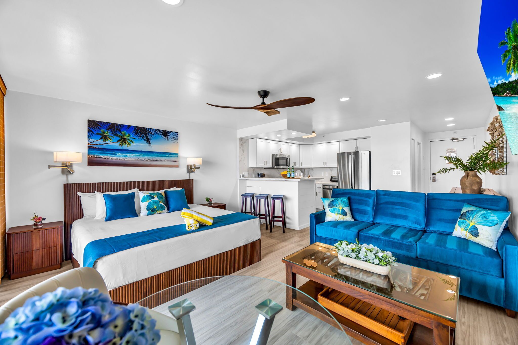 A modern hotel room with a bed, a blue sofa, a glass coffee table, a kitchen area, and a ceiling fan.