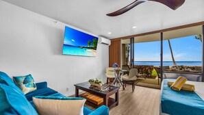 A modern living room with a large TV, a sofa, a coffee table, and a balcony with a view of the ocean.