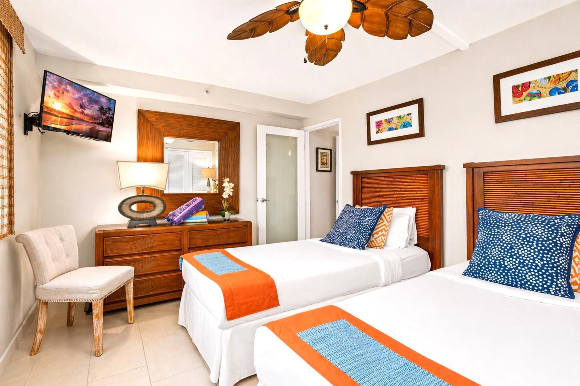 Quarto de hotel com duas camas, uma escrivaninha com abajur, uma cadeira, ventoinha no teto e obras de arte nas paredes.