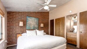 A bedroom with a brick wall, a bed with white linens, a ceiling fan, and a wooden dresser with a mirror. - Struggler 3 Bedrooms STR-1 by KBM (Park City)