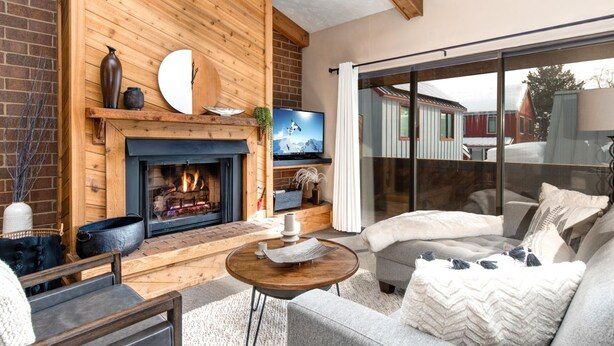 A cozy living room with a fireplace, a television, and a large sliding glass door leading to an outdoor area. - Struggler 3 Bedrooms STR-1 by KBM (Park City)