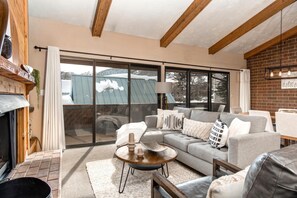 A modern living room with a grey sofa, a round coffee table, and a fireplace. Large windows offer a view of a snowy landscape. - Struggler 3 Bedrooms STR-1 by KBM (Park City)