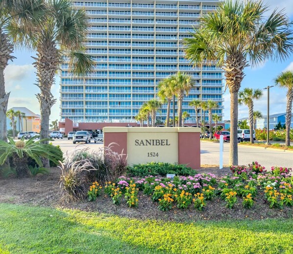 L’entrée d’un hôtel, avec une enseigne indiquant « SANIBEL 1524 », est bordée d’un jardin aménagé où poussent différentes variétés de fleurs et de palmiers.