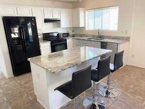 A kitchen with white cabinets, a black refrigerator, a stove, a microwave, and a sink. There are four bar stools around the island. - Minutes from the mountains and the Rillito River (Tucson)