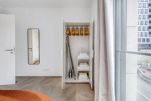 Une chambre d’hôtel moderne dotée d’une grande fenêtre, d’une armoire où sont suspendus des vêtements et d’un miroir au mur.