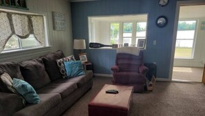Una sala de estar con sofá, sillón y reposapiés, una ventana con cortinas y una puerta que da al exterior.