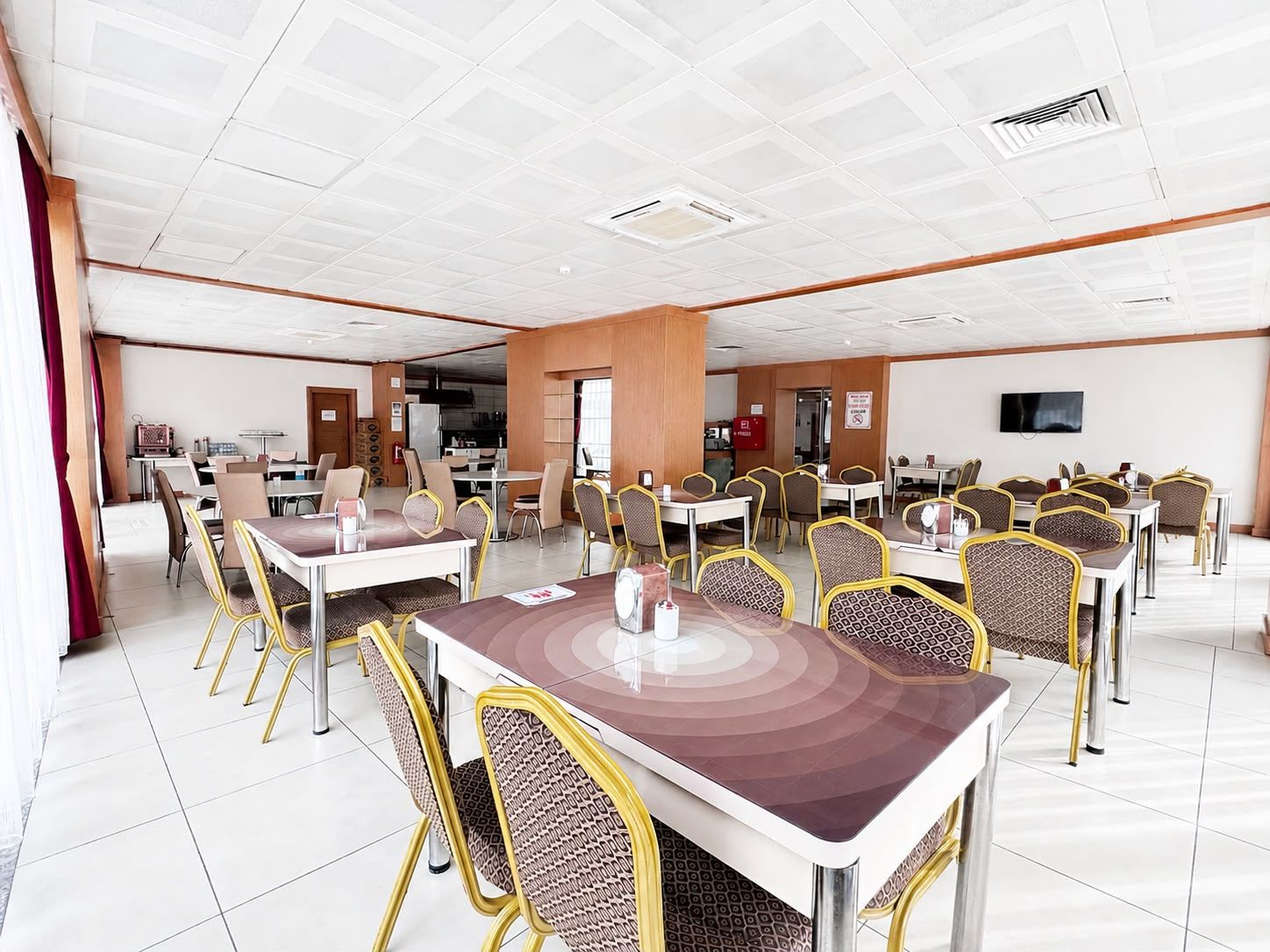 A spacious dining area with multiple tables and chairs, a television mounted on the wall, and a kitchen area in the background.