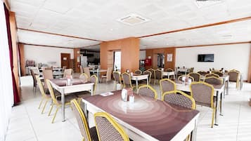 A spacious dining area with multiple tables and chairs, a television mounted on the wall, and a kitchen area in the background.