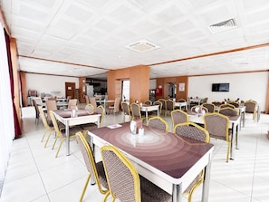 A spacious dining area with multiple tables and chairs, a television mounted on the wall, and a kitchen area in the background. - Quars Garden Hotel (Düzce)
