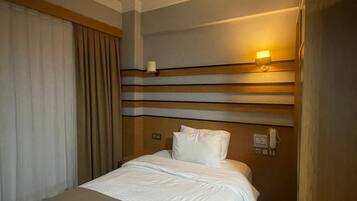 Una habitación de hotel con una cama, un ventilador de techo, una lámpara fijada a la pared y una ventana con cortinas.