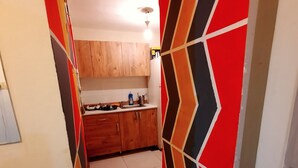 A narrow hallway with a kitchen area at the end. The hallway has a geometric patterned wall on one side and a plain wall on the other. The kitchen has wooden cabinets and a white countertop. - Lovely 1-Bed Apartment (Istanbul)