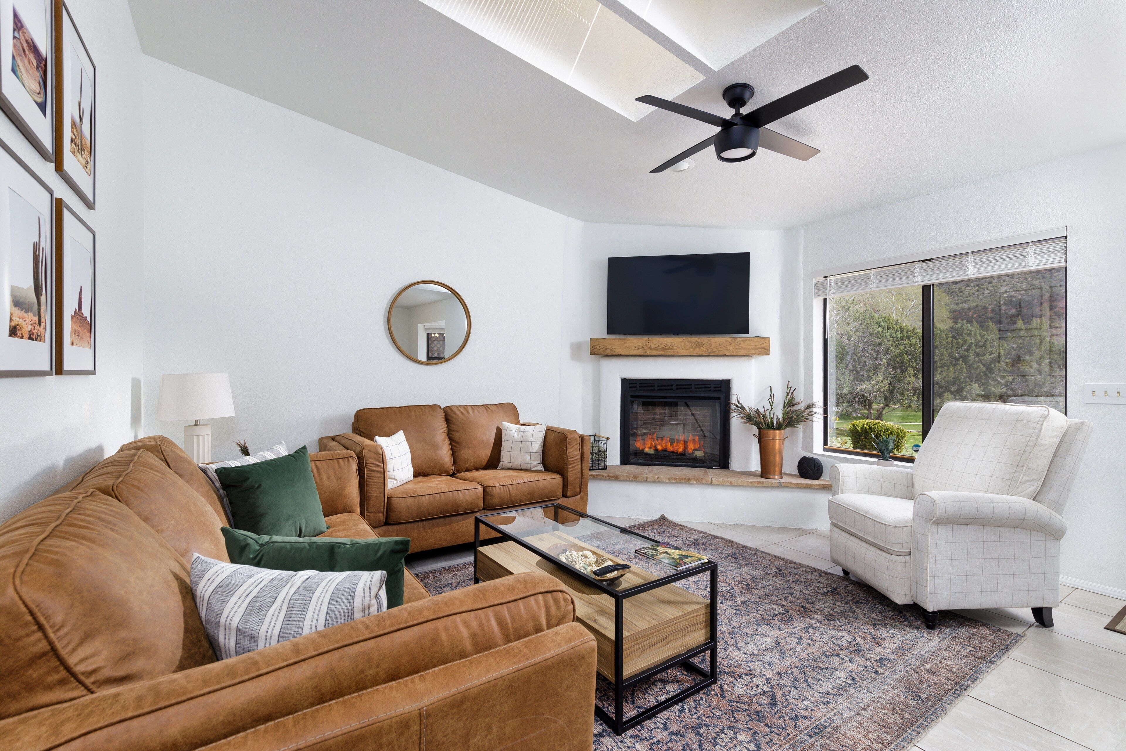 A modern living room with a brown leather sofa, a white armchair, a glass coffee table, a fireplace, and a large window.