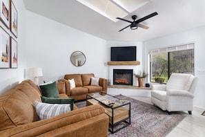 A modern living room with a brown leather sofa, a white armchair, a glass coffee table, a fireplace, and a large window.