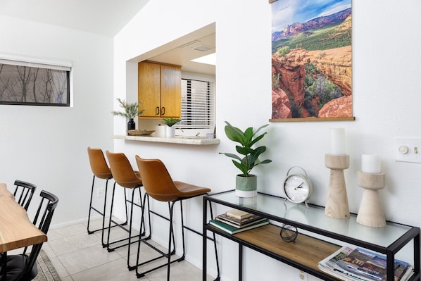 A modern kitchen with a dining area, featuring a wooden table, bar stools, a small island with a potted plant, and a wall-mounted clock.