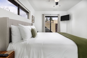 A bedroom with a large bed, a TV mounted on the wall, and a view of the outside through a sliding door.