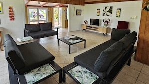 A spacious living area with a large sofa set, a coffee table, and a TV. There are wooden beams on the ceiling and a hanging light fixture. - Ann's Beach House (Rarotonga)