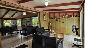 A spacious living area with a sofa, chairs, and a dining table. There is a fan and a decorative cabinet with various items. - Ann's Beach House (Rarotonga)