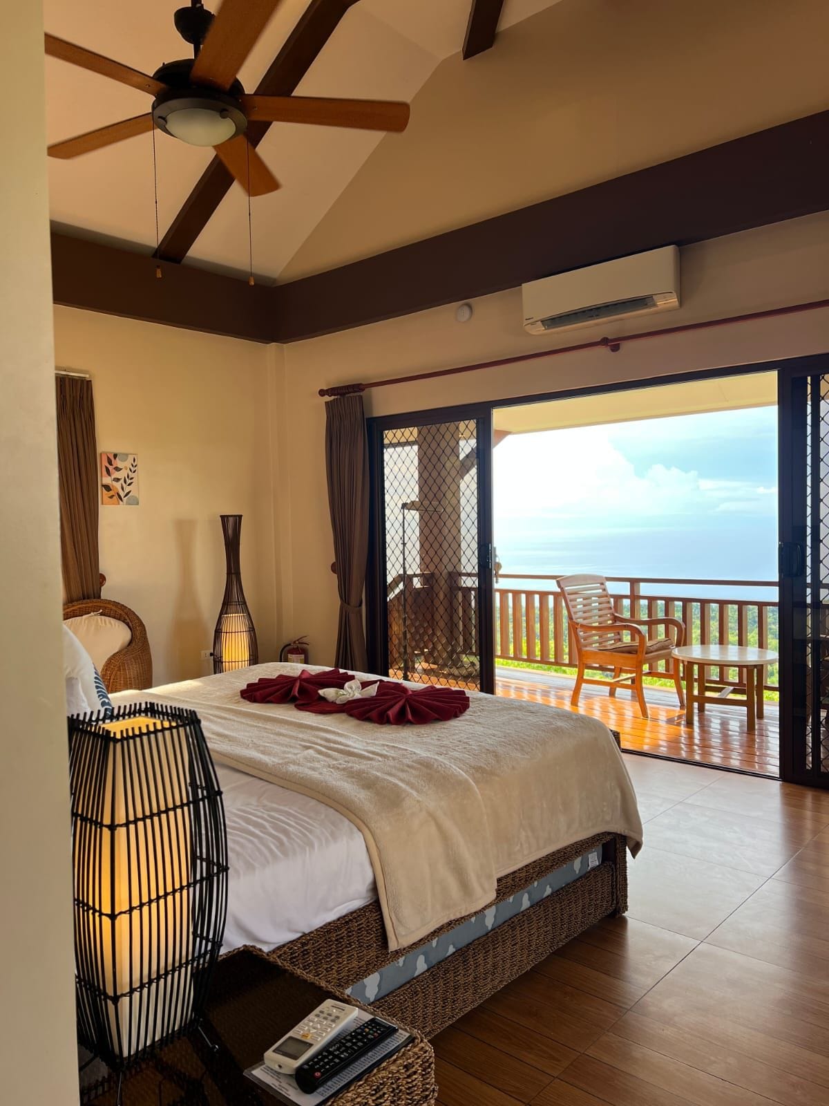Ein Schlafzimmer mit Bett, Deckenventilator, Nachttisch, Lampe und Blick auf den Ozean vom Balkon aus.