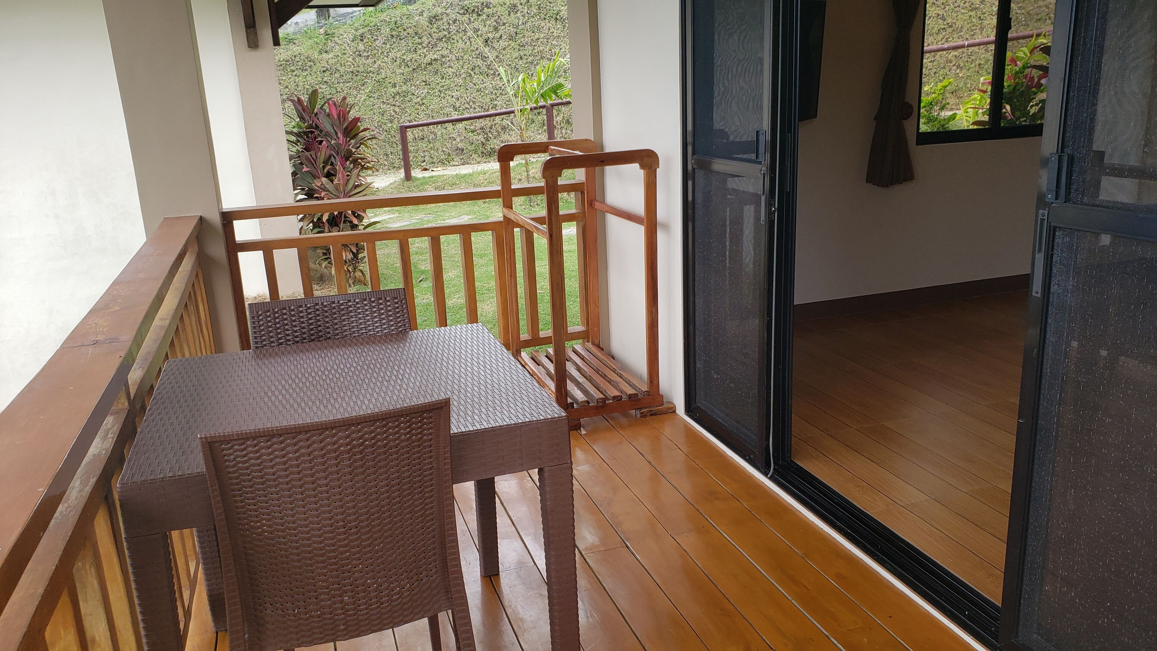 A balcony with a wooden table and chairs, a view of a garden, and a sliding glass door.