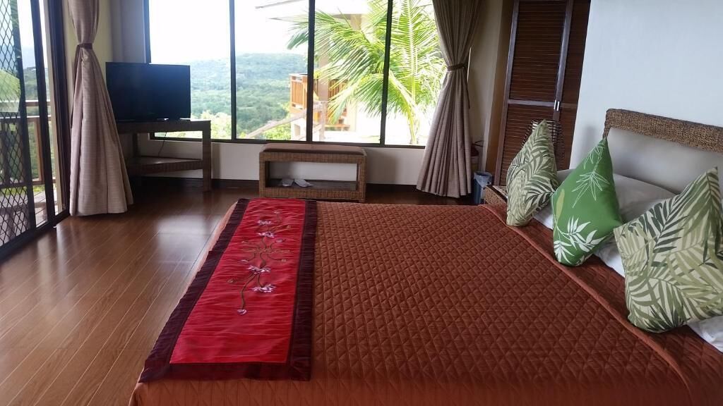 Un dormitorio con una cama grande, un televisor sobre un soporte, piso de madera y vista a palmeras por la ventana.