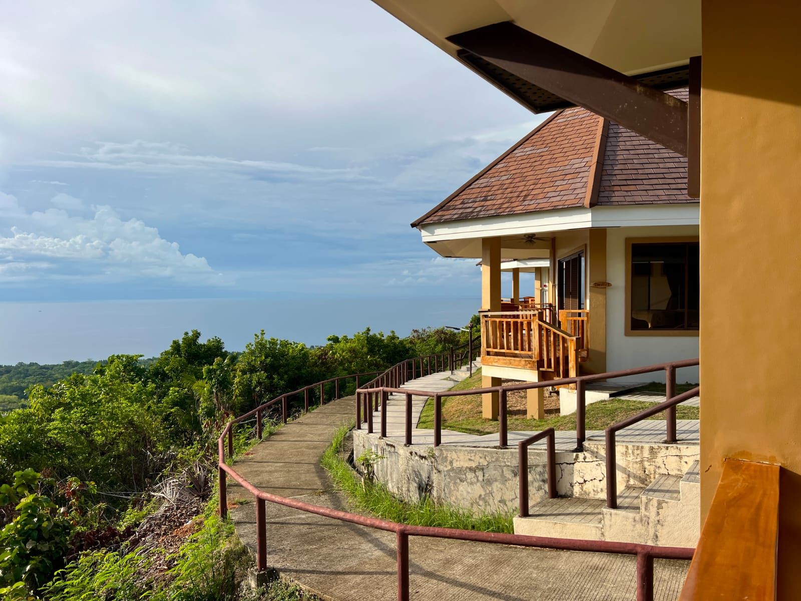 A coastal property with a balcony overlooking the sea, a concrete pathway leading to the entrance, and lush greenery surrounding the area.