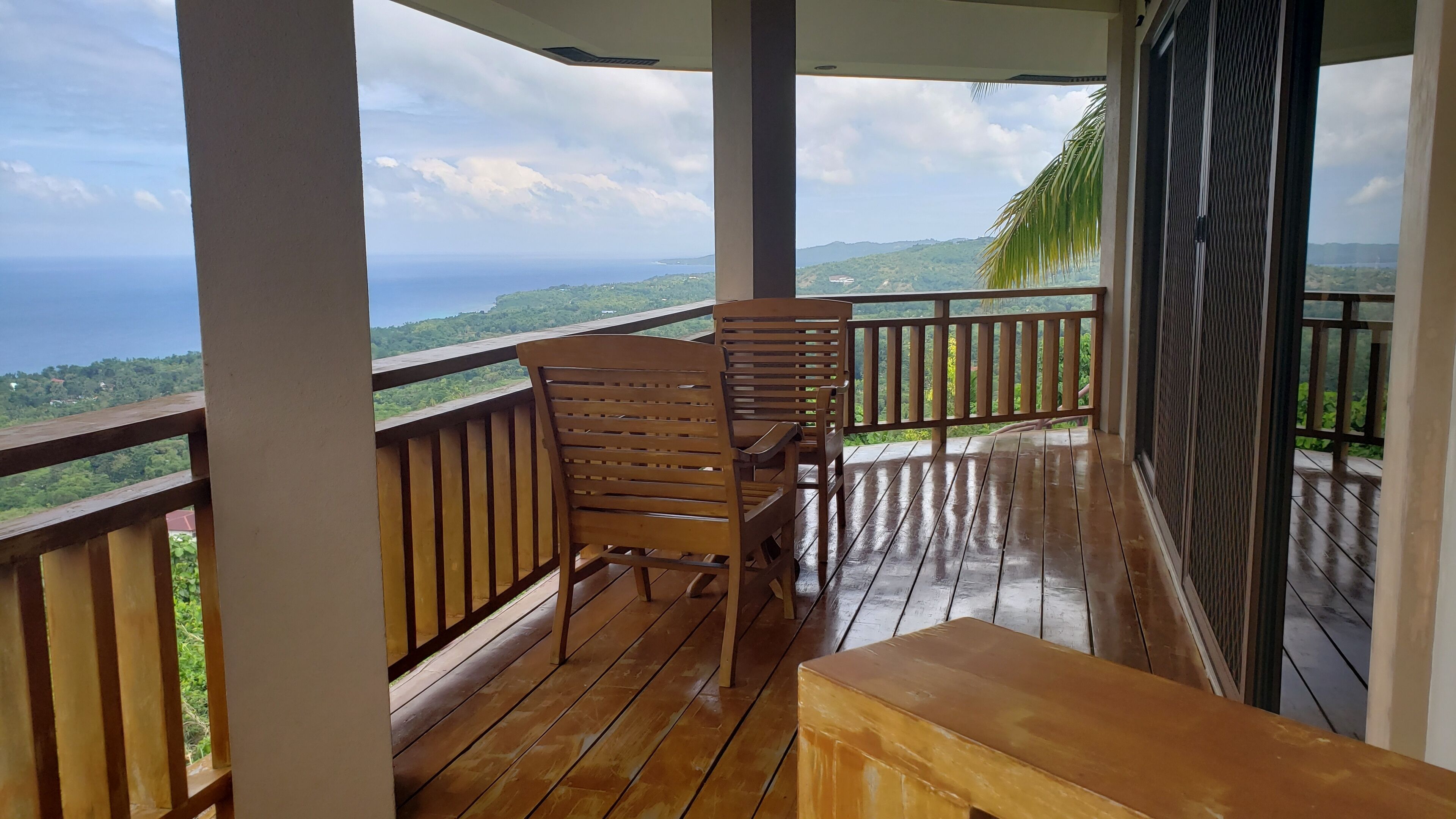 A wooden deck with a bench and chairs, overlooking a scenic view of the ocean and hills.