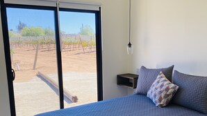 A bedroom with a large bed, a nightstand, a hanging light, and a sliding glass door leading to an outdoor area with a vineyard.