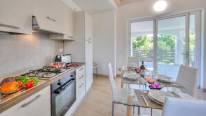 A modern kitchen with a gas stove, oven, and white cabinets. There is a dining area with a glass table and chairs, and a view of greenery outside. - Golden Terrace Leggiuno 2BR/2BA Walk to the Lake, Leggiuno, Italy (Leggiuno)