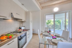 Cozinha moderna com fogão a gás, forno e armários brancos. Há uma área de jantar com mesa e cadeiras de vidro, e vista para o verde lá fora.