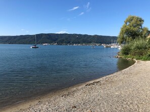 Un lac paisible où flottent des bateaux, une plage de galets et, au loin, une colline boisée.