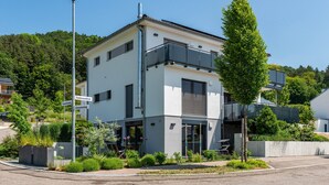 Moderni, kaksikerroksinen rakennus, jossa on parveke, ja sen ympärillä on vehreää kasvillisuutta ja avoin, kirkas taivas.