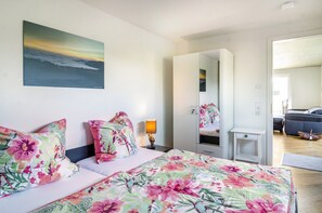 Une chambre à coucher avec un lit, une literie à motifs floraux, une table de chevet avec une lampe, un grand miroir et une vue sur une autre pièce.