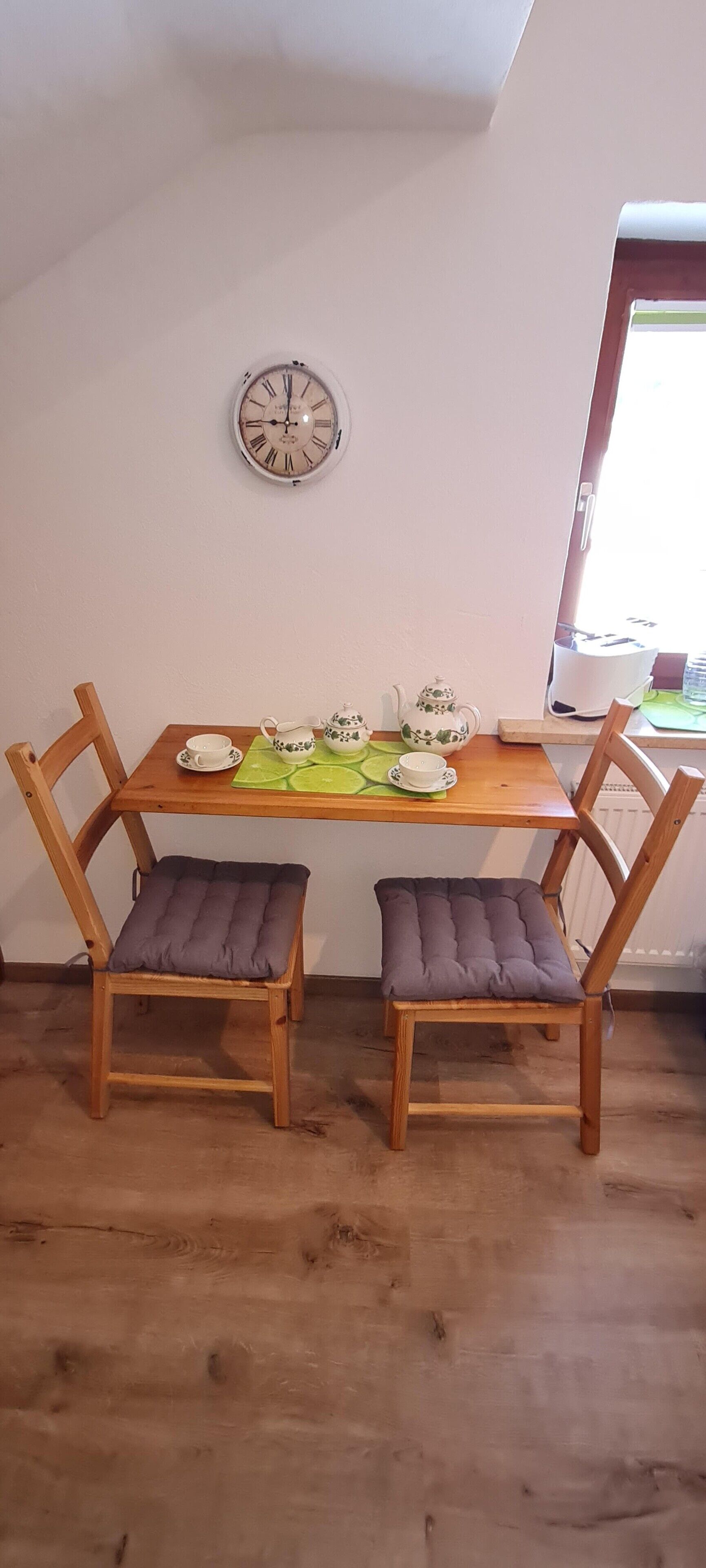 Een eethoek met een houten tafel, twee stoelen, een klok en een raam.