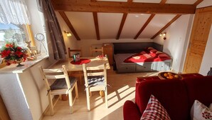 A cozy room with a wooden ceiling, a bed with a red blanket, a red sofa, a dining table with chairs, and a window with curtains. - Apartment 'Haus Bergblick' with Balcony and Wi-Fi (Mittenwald)
