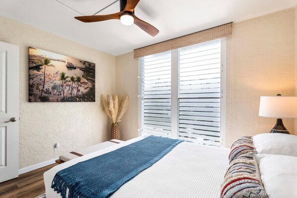 Une chambre à coucher comprenant un lit, un ventilateur de plafond, une fenêtre avec des stores, un tableau accroché au mur et une lampe de chevet.